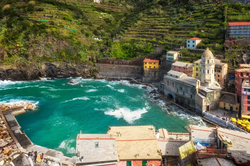 Vista di Vernazza nelle Cinque Terre con mare turchese, case colorate e terrazze verdi.